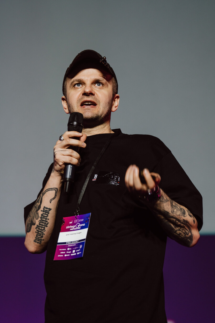 Osoba z tatuażami na obu ramionach, ubrana w czarną koszulę i czapkę, mówi do mikrofonu na scenie. Na szyi ma plakietkę konferencji i gestykuluje drugą ręką.