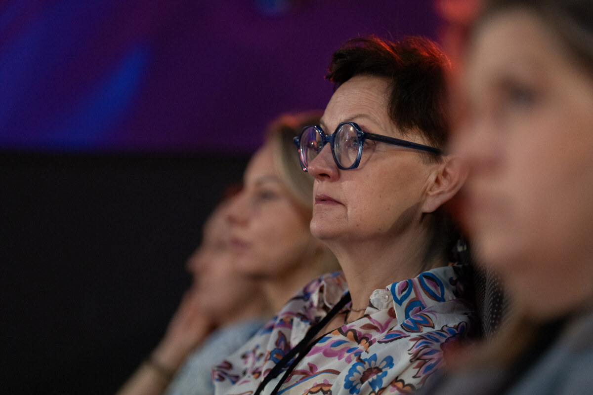 Kobieta z krótkimi włosami i niebieskimi okularami, ubrana w kolorową kwiecistą koszulę, uważnie słucha na widowni. Dwie inne kobiety są widoczne w miękkiej ostrości obok niej. Tło jest ciemne i rozmyte.