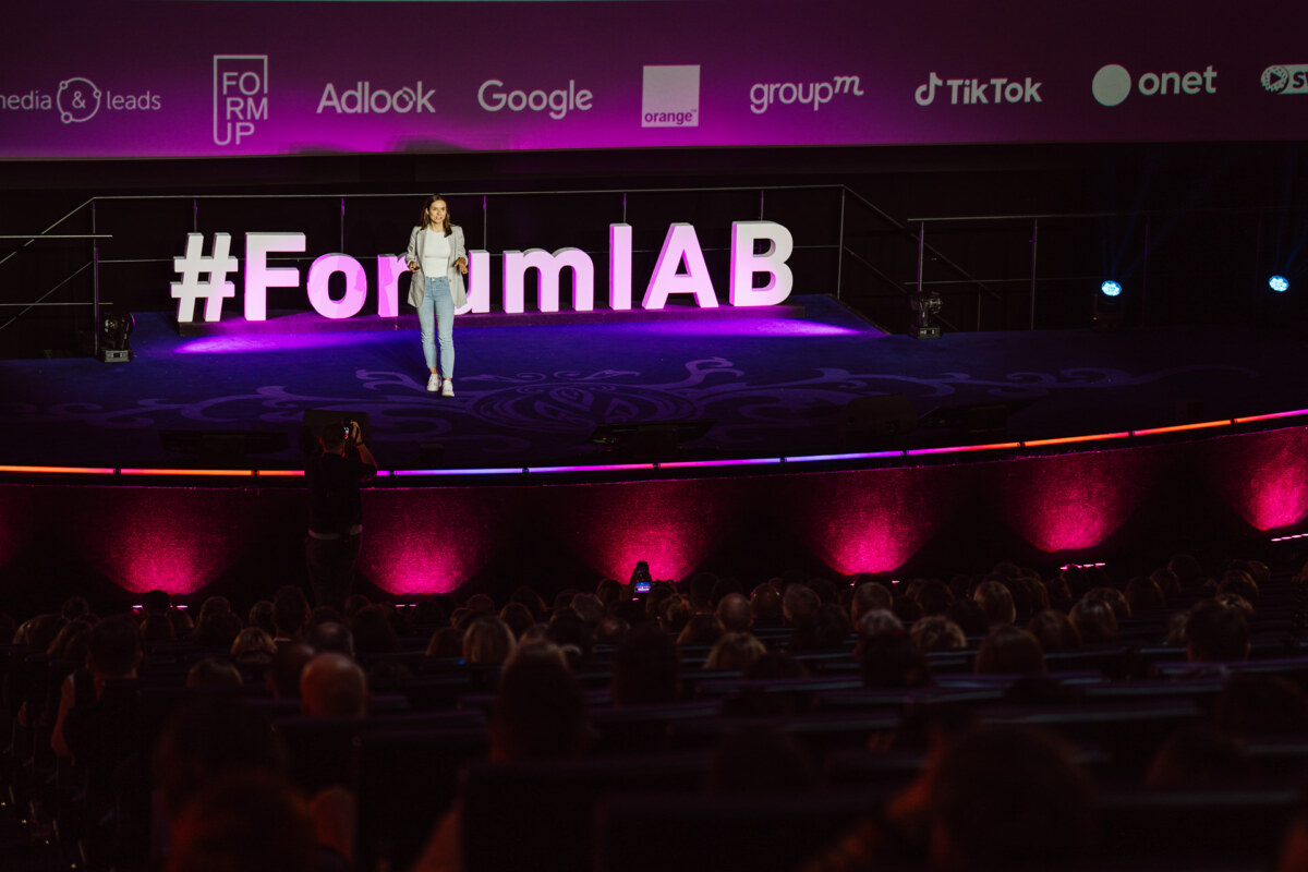 Kobieta stoi na scenie przemawiając na konferencji z dużym napisem "#ForumIAB" za nią i ekranem pokazującym logo sponsorów. Publiczność siedzi przed sceną.