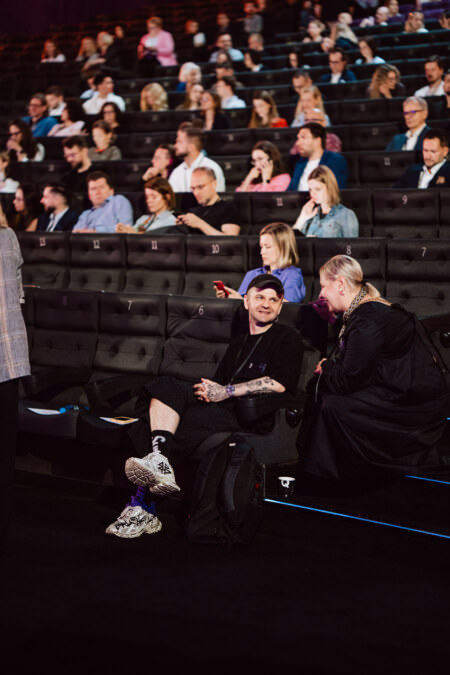 Ein Mann und eine Frau sitzen in der ersten Reihe eines vollbesetzten Theaters und unterhalten sich. Der Mann trägt eine blaue Mütze und Turnschuhe, während die Frau kurzes blondes Haar und einen langen Mantel trägt. Das Publikum sitzt auf nummerierten Plätzen hinter ihnen.  