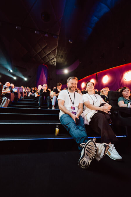 Ein Mann und eine Frau sitzen lächelnd auf den dunklen Stufen eines Theaters inmitten einer von farbigen Lichtern erleuchteten Menge. Die Beine des Mannes sind ausgestreckt und beide tragen Event-Lanyards. Die Atmosphäre ist lebhaft und entspannt.  