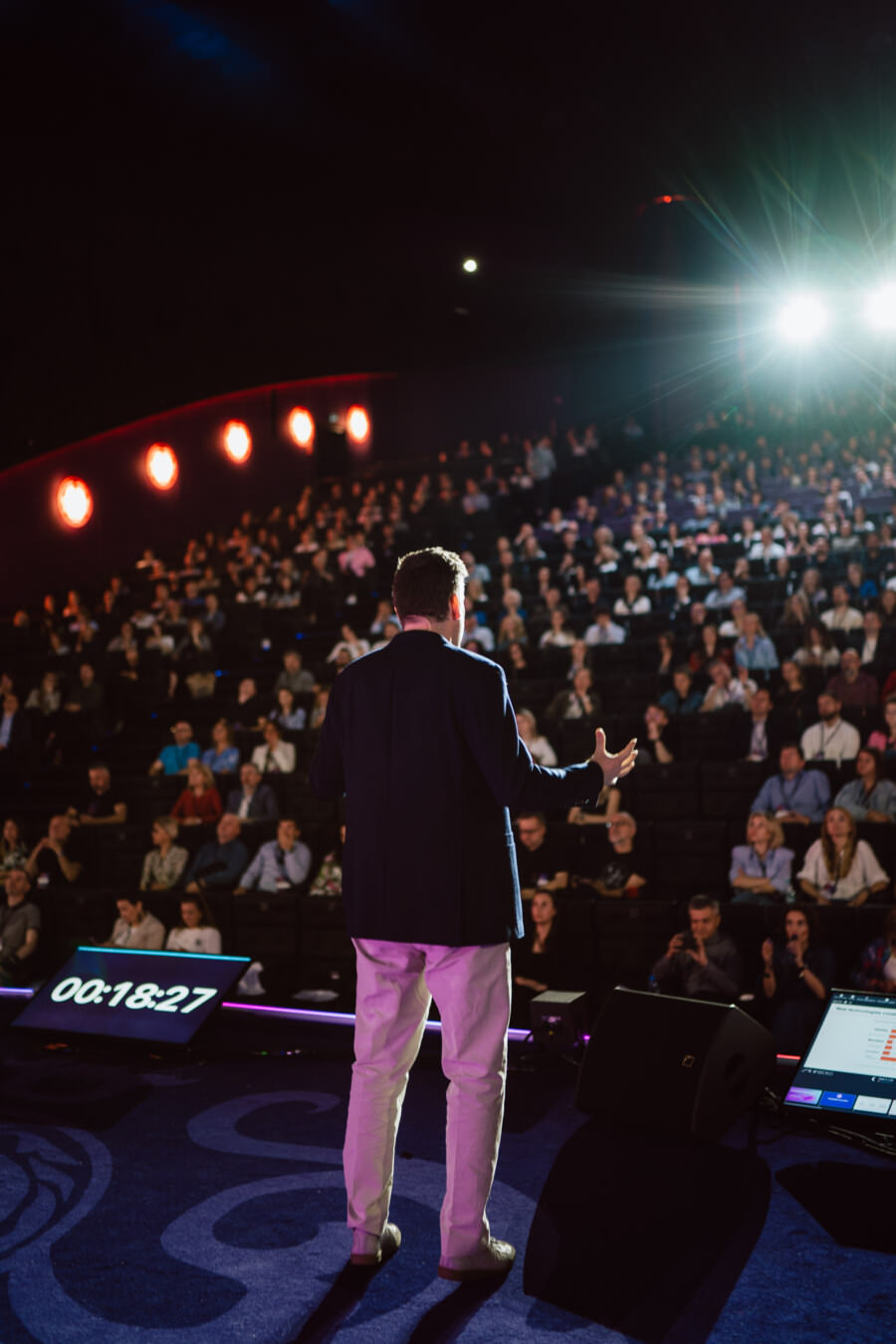 Eine Person steht auf einer Bühne vor einem sitzenden Publikum in einem dunklen Auditorium und spricht bei hellem Licht. Zwei große Digitaluhren sind auf dem Boden der Bühne zu sehen. Das Publikum scheint aufmerksam zu sein.  