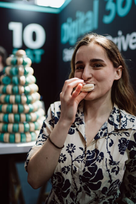 Eine junge Frau in einem geblümten Hemd lächelt, während sie ein Macaron isst. Im Hintergrund sehen Sie eine große Pyramide aus Macarons und einen unscharfen digitalen Hintergrund.