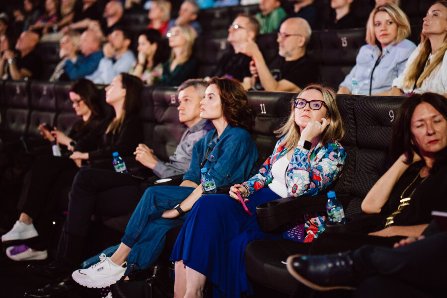 Publiczność siedzi w słabo oświetlonym teatrze i uważnie obserwuje. Ludzie w różnym wieku wydają się skupieni, niektórzy z zamyślonymi wyrazami twarzy. Kobieta na pierwszym planie nosi okulary i kolorową kurtkę, opierając podbródek na dłoni.