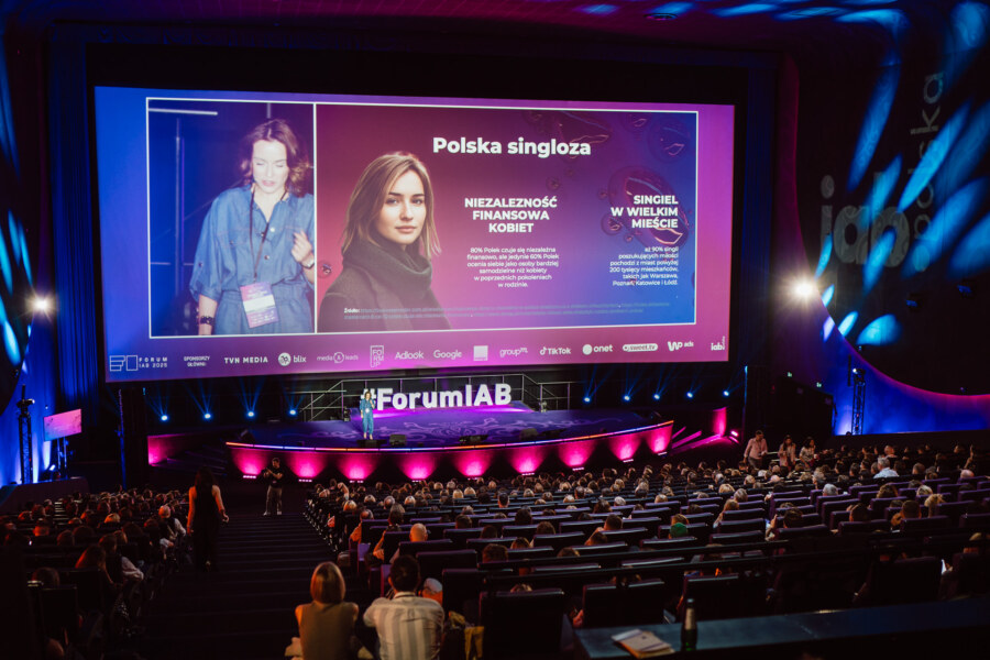 Kobieta stoi na scenie podczas konferencji w dużym audytorium. Za nią znajduje się duży ekran wyświetlający slajdy w języku polskim na temat niezależności finansowej kobiet. Uczestnicy siedzą i oglądają prezentację.