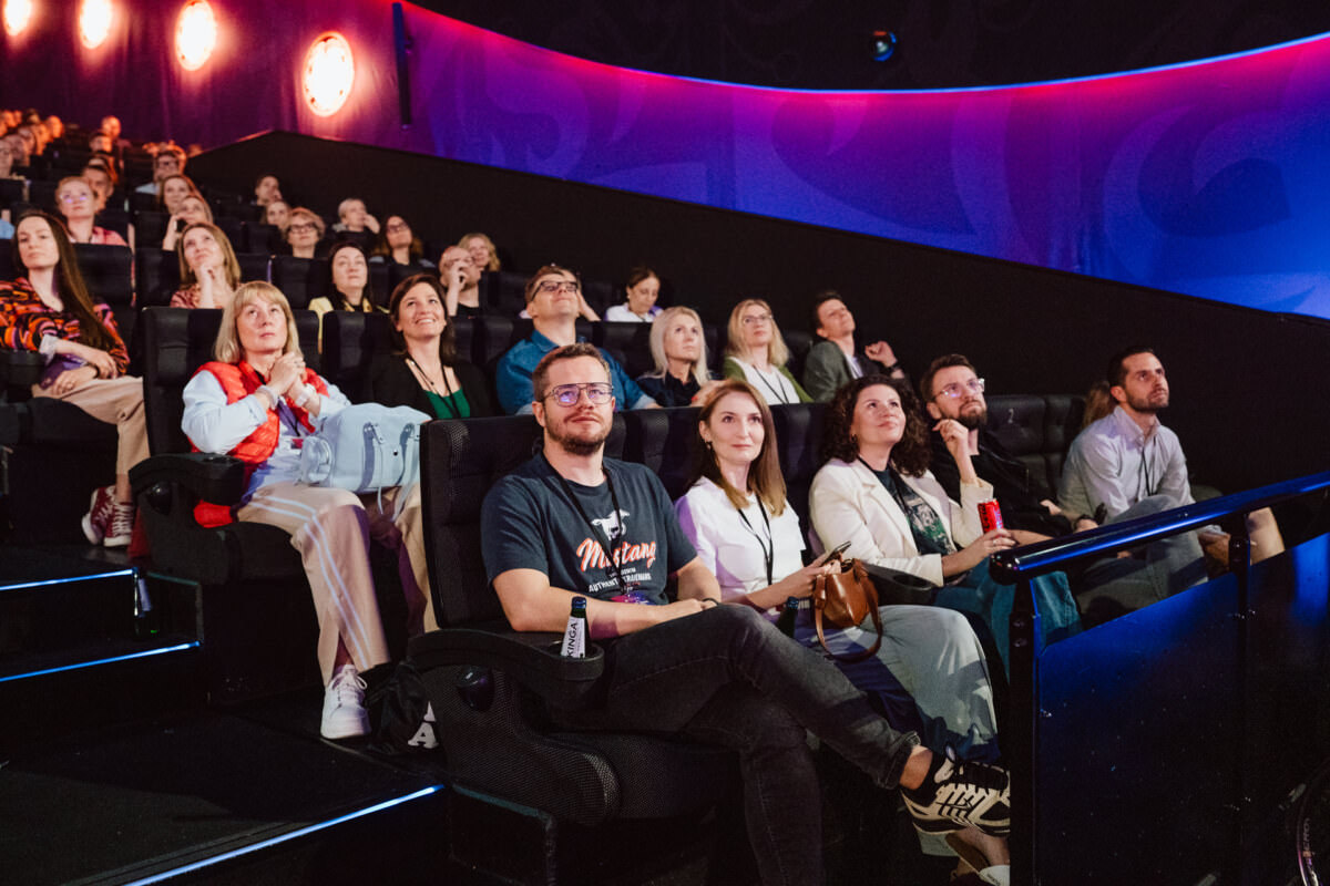 Eine bunt gemischte Gruppe von Menschen sitzt in einem dunklen Theater und schaut aufmerksam auf die Leinwand. Der Raum wird von farbigen Lampen beleuchtet und das Publikum scheint engagiert und konzentriert zu sein. 