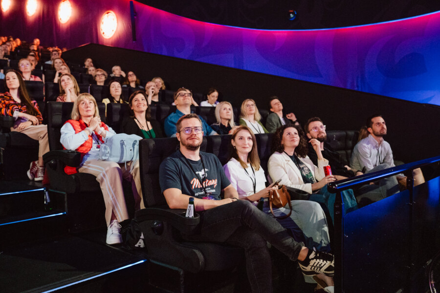 Zróżnicowana grupa ludzi siedzi w ciemnym teatrze, uważnie obserwując ekran. Pokój jest oświetlony kolorowymi kinkietami, a publiczność wydaje się zaangażowana i skupiona.