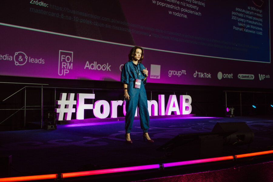 Kobieta w niebieskim kombinezonie stoi na scenie przemawiając na konferencji, z dużym napisem #ForumIAB i kolorowymi logo sponsorów wyświetlanymi za nią.