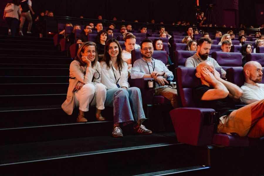 Grupa ludzi siedzi w teatrze z fioletowymi siedzeniami, niektórzy uśmiechają się do kamery, podczas gdy inni patrzą przed siebie. Trzy osoby w pierwszym rzędzie siedzą na schodach, wyglądając na zrelaksowane i wesołe.
