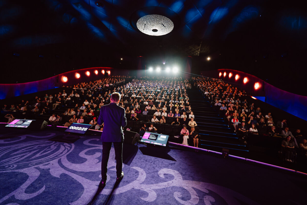 Der Redner steht auf der Bühne vor einem großen Publikum, das in einem modernen Auditorium sitzt, das von Bühnenscheinwerfern und kräftiger blauer und roter Beleuchtung erhellt wird. An den Rändern der Bühne sind Präsentationsbildschirme zu sehen. 