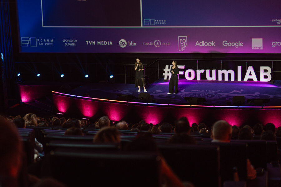 Dwie osoby stoją na scenie, prowadząc prezentację podczas wydarzenia Forum IAB, z dużą publicznością siedzącą z przodu. Na tle wyświetlane są logotypy sponsorów i marki wydarzenia w odcieniach fioletu i niebieskiego.