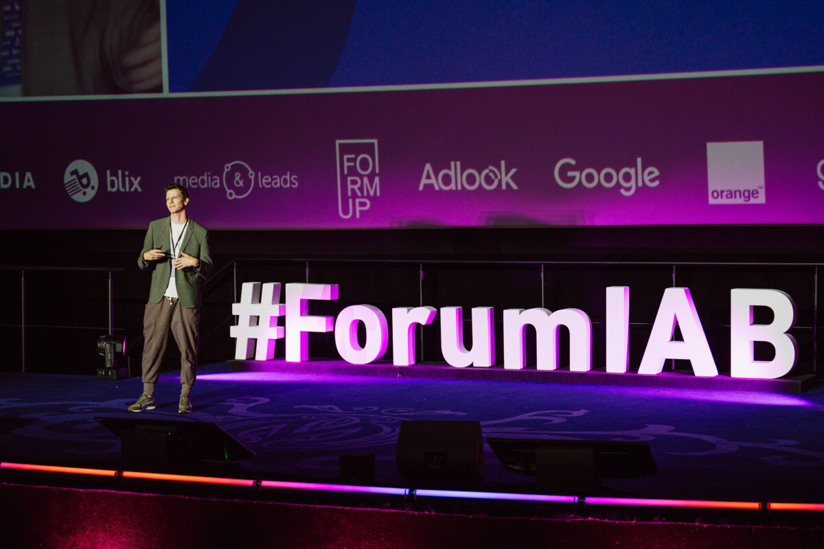 Mężczyzna w stroju biznesowym stoi na scenie obok dużych podświetlanych liter z napisem "#ForumIAB", z logo firmy i fioletowym tłem wyświetlanym za nim.