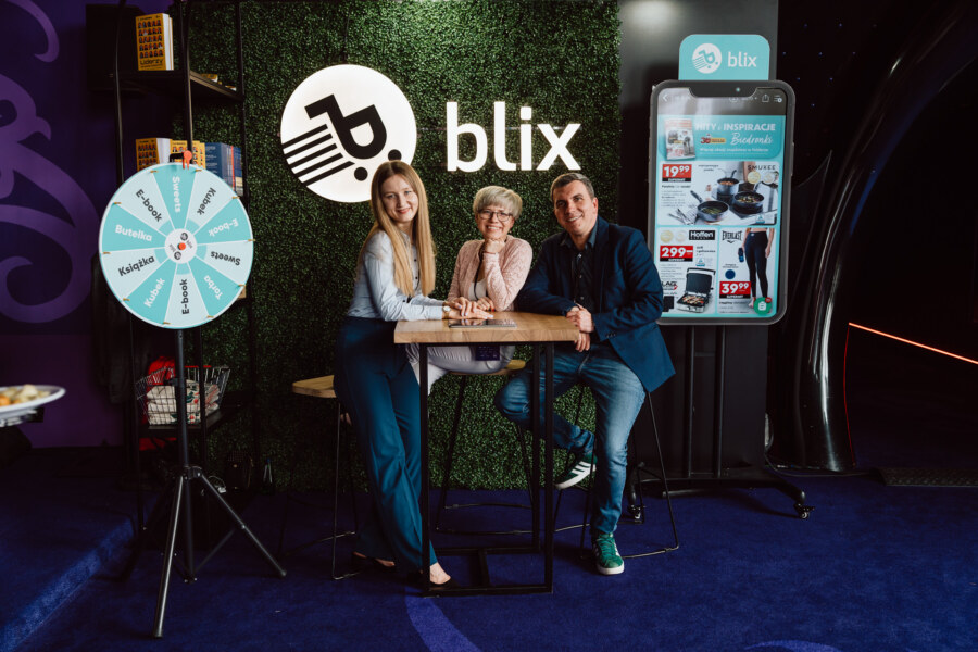 Trzy osoby stoją i siedzą wokół wysokiego stołu przed zieloną ścianą z logo "blix". W pobliżu znajduje się obracające się koło nagród i stoisko promocyjne. Wszystkie trzy osoby są uśmiechnięte i zwrócone w stronę kamery.