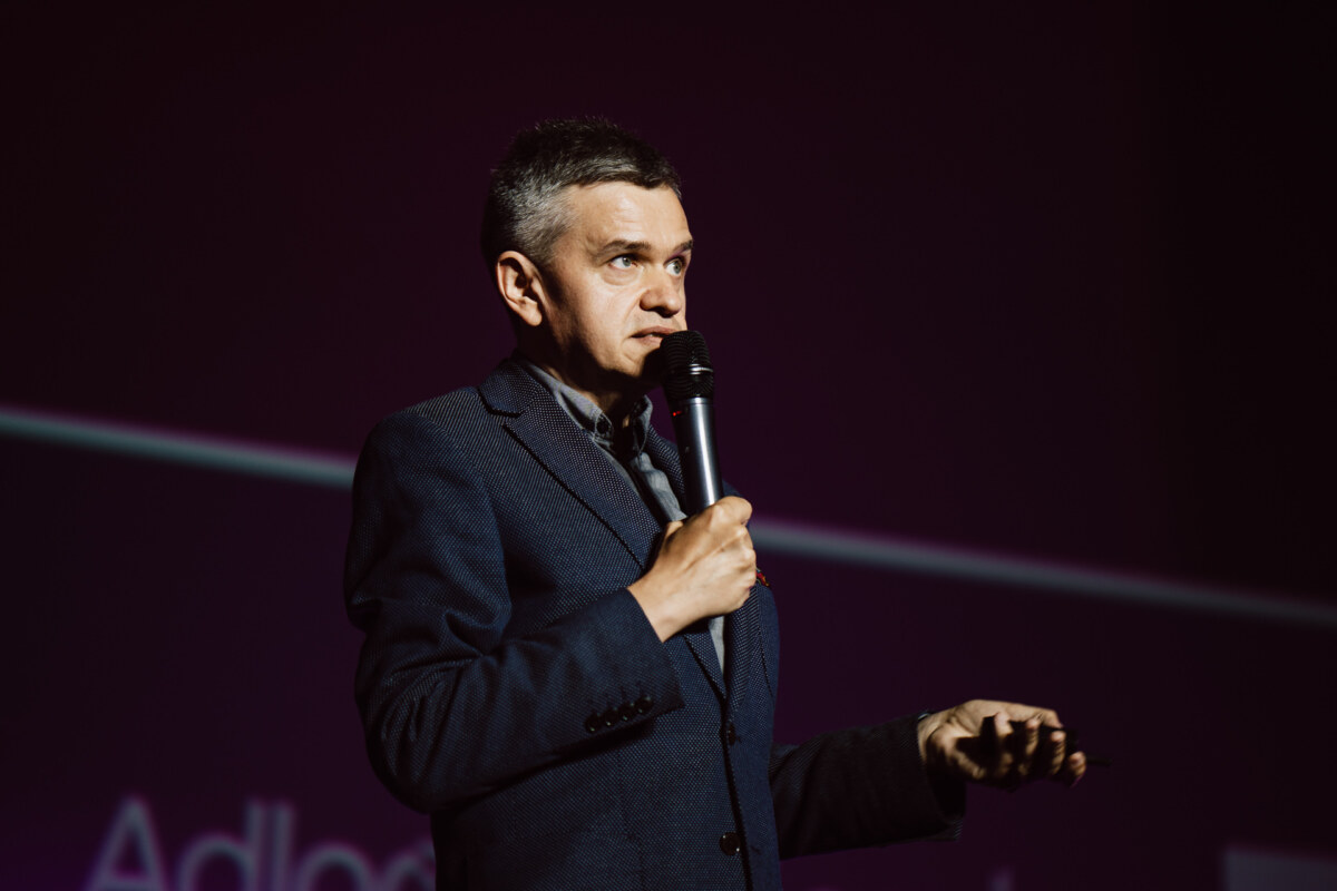 Mężczyzna w ciemnym garniturze trzyma mikrofon i przemawia na scenie na fioletowym tle, wyglądając na zaangażowanego i skupionego podczas swojej prezentacji.