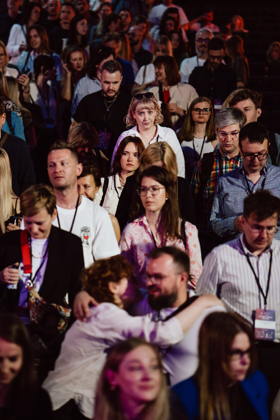 Duża grupa ludzi stoi blisko siebie na imprezie wewnętrznej, wielu z nich nosi smycze. Niektórzy patrzą bezpośrednio w kamerę, podczas gdy inni rozmawiają, chodzą lub obejmują się. Oświetlenie jest przyćmione z kolorowymi refleksami.