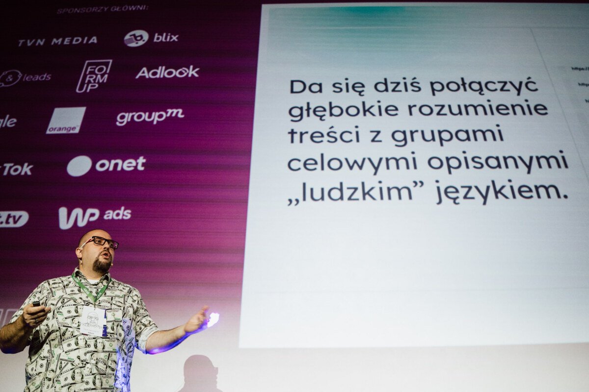 Mężczyzna stoi na scenie przed ekranem, na którym wyświetlany jest polski tekst i logotypy sponsorów. Slajd omawia połączenie głębokiego zrozumienia treści z grupami docelowymi za pomocą "ludzkiego" języka.