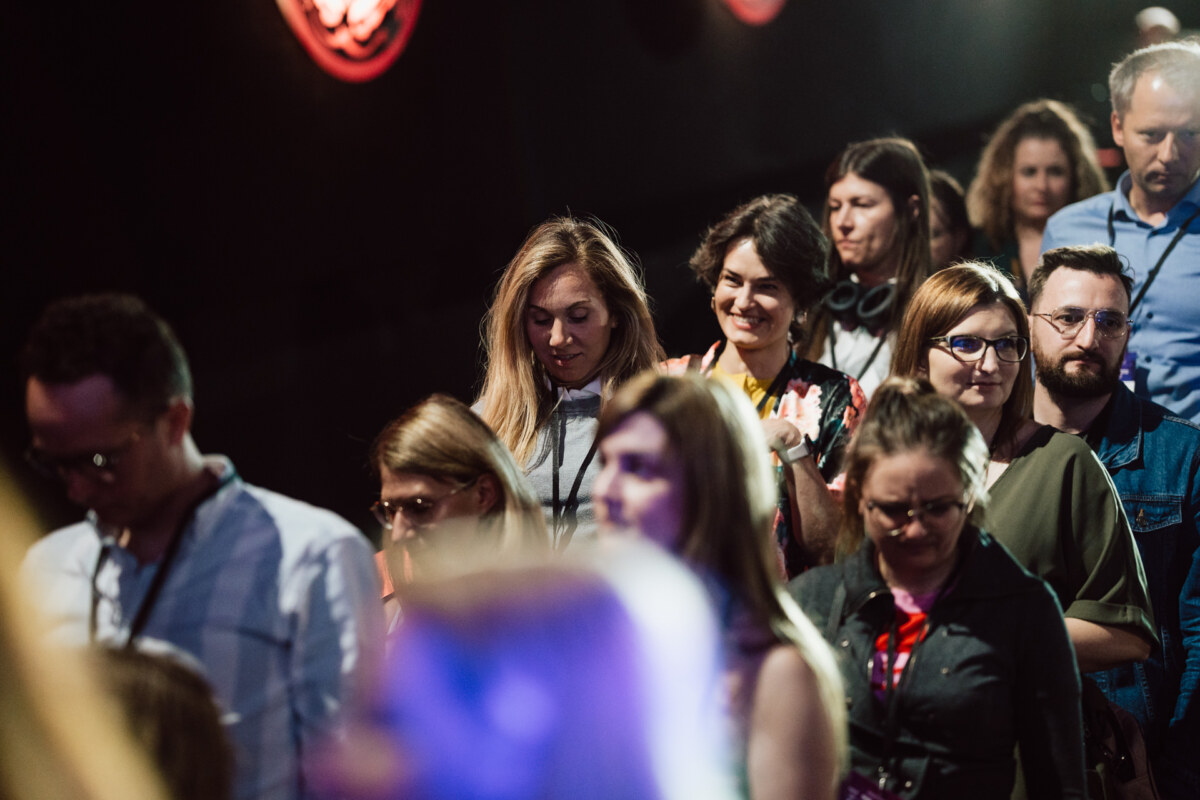 Grupa ludzi, zarówno mężczyzn, jak i kobiet, idzie blisko siebie w pomieszczeniu, niektórzy patrzą w dół, podczas gdy inni się uśmiechają. Większość z nich ma na sobie identyfikatory konferencyjne i swobodne ubrania w słabo oświetlonym otoczeniu.