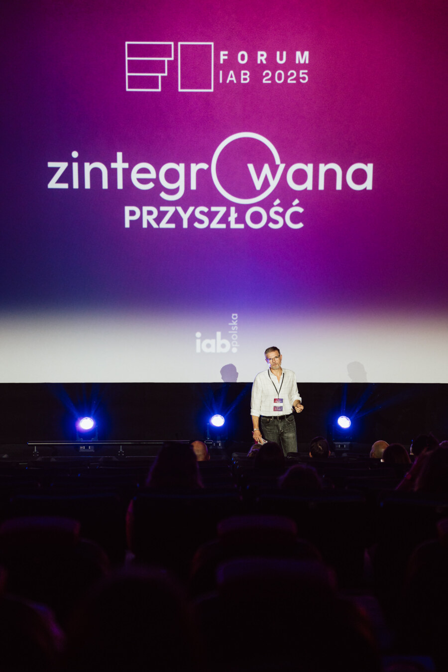 Prelegent stoi na scenie przed dużym ekranem wyświetlającym "Forum IAB 2025" i "zintegrowaną przyszłość" dla publiczności w ciemnym teatrze.