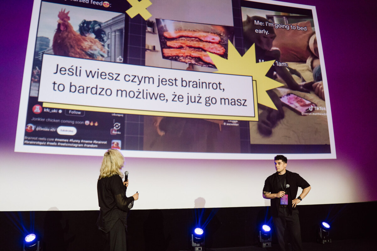 Dwie osoby stoją na scenie z mikrofonami, naprzeciwko dużego ekranu wyświetlającego polski tekst i zdjęcia jedzenia i zwierząt z mediów społecznościowych. Niebieskie światła sceniczne oświetlają scenę.
