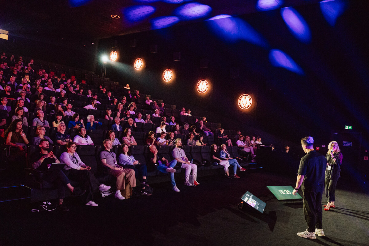 Dwóch prezenterów stoi na scenie naprzeciwko siedzącej publiczności w słabo oświetlonym teatrze. Publiczność przygląda się uważnie, a okrągłe światła zdobią ciemne ściany. Niebieskie i fioletowe światła sceniczne podkreślają scenę.