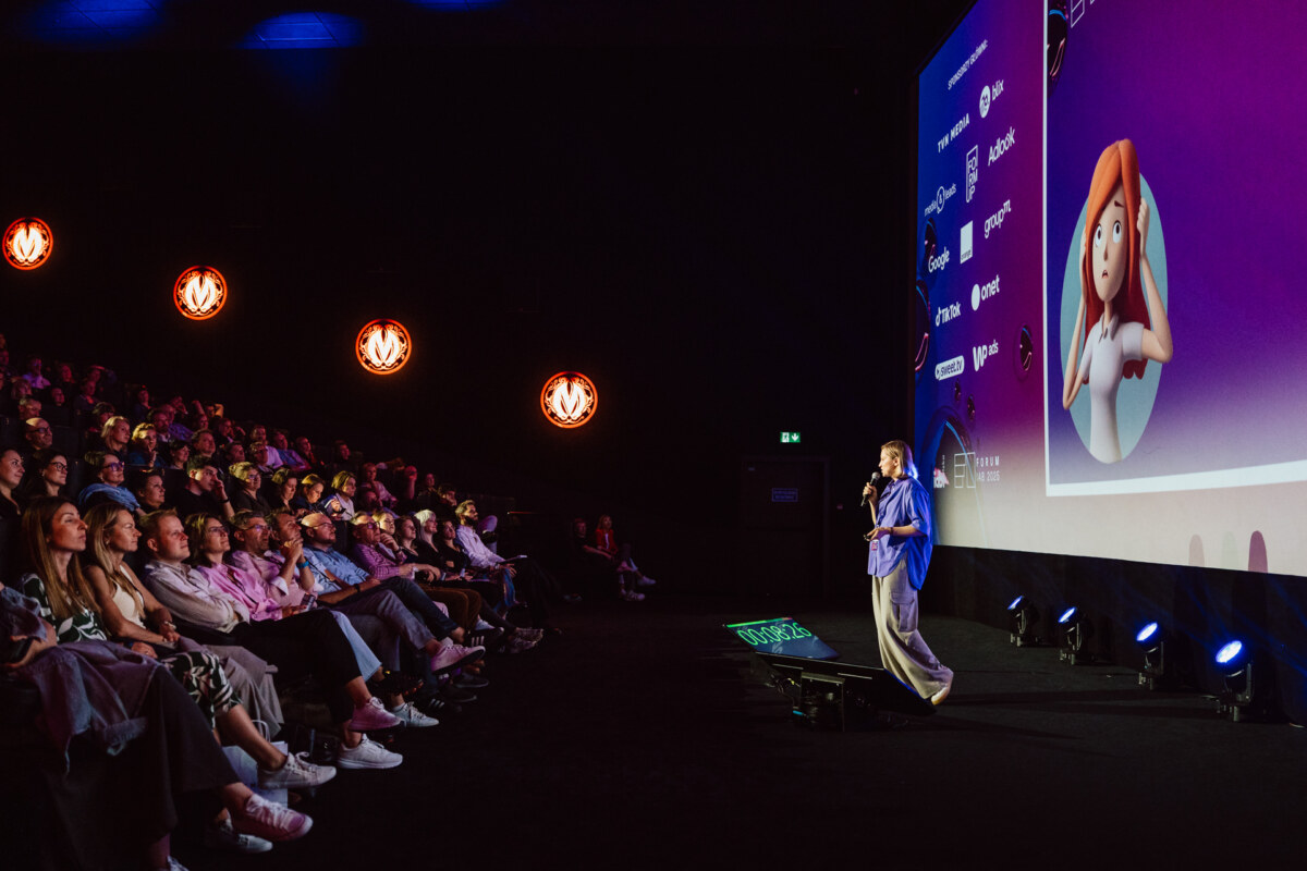 Prelegent stoi na scenie, prezentując przed dużą publicznością siedzącą w ciemnym teatrze, z wyświetlanym ekranem wyświetlającym logo i postać z kreskówki. Pomieszczenie jest oświetlone pomarańczowymi kinkietami.