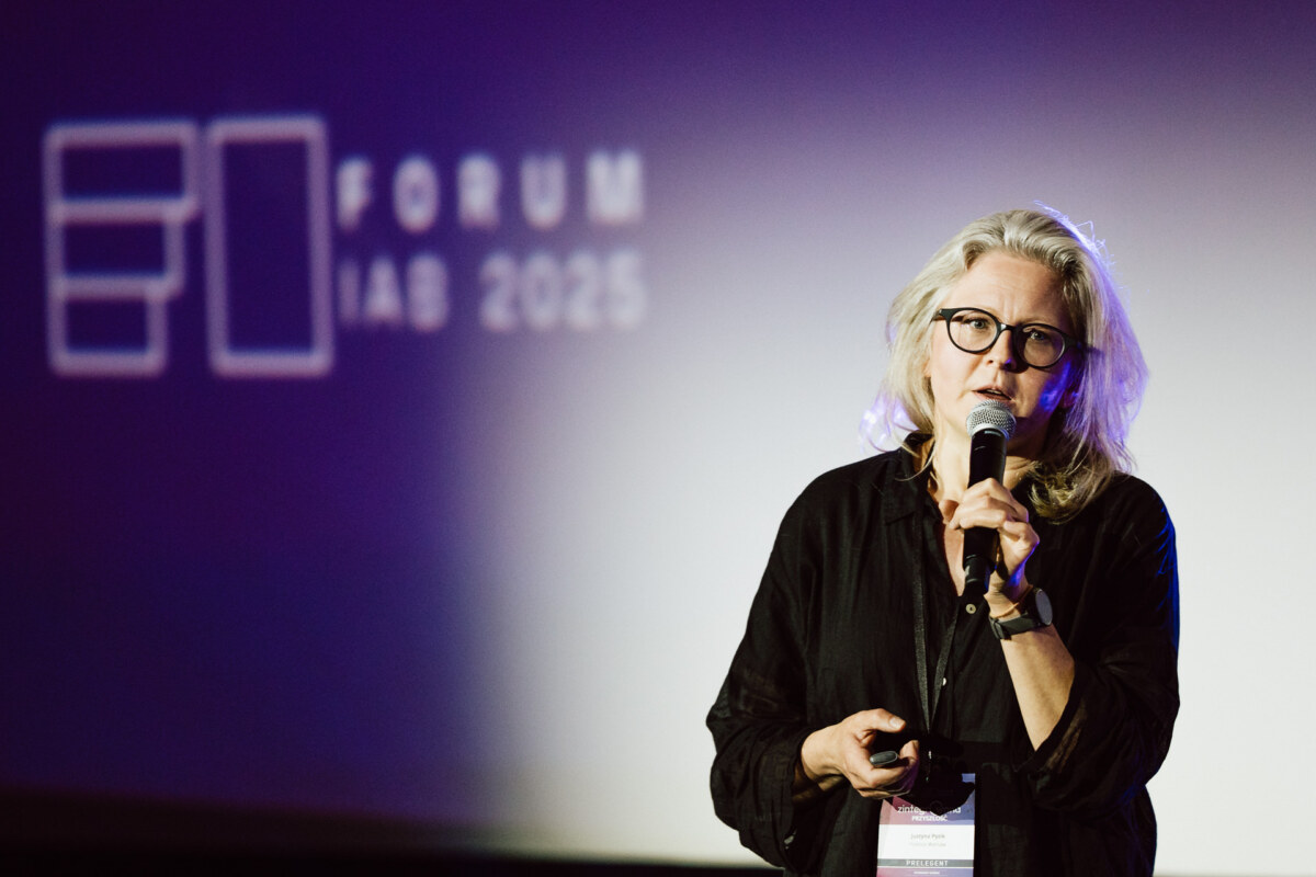 Kobieta w okularach i siwych włosach mówi do mikrofonu na scenie, trzymając pilota. Za nią na ekranie wyświetla się napis "FORUM LAB 2025".