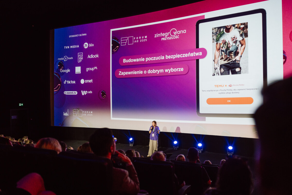 Kobieta przemawia na scenie w ciemnym audytorium przed dużym ekranem wyświetlającym polski tekst, cyfrową grafikę i logo sponsorów. Publiczność siedzi w rzędach, oglądając prezentację.