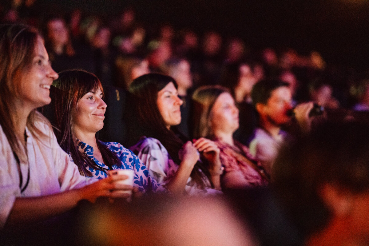 Grupa ludzi siedzi blisko siebie w słabo oświetlonym teatrze, uśmiechając się i oglądając coś na scenie lub ekranie. Publiczność wygląda na zaangażowaną i rozbawioną.