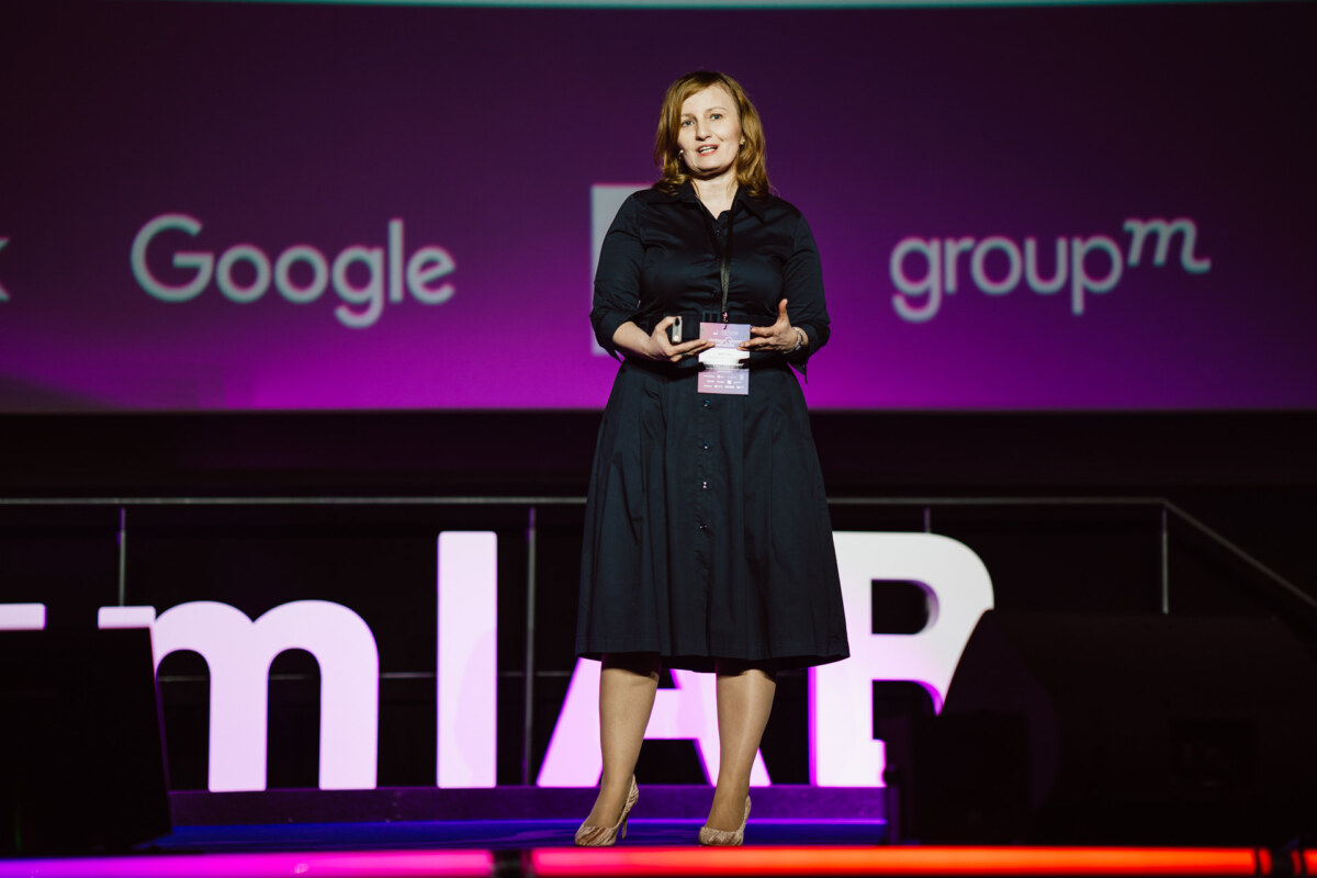 Kobieta w ciemnej sukience stoi boso na scenie, przemawiając do publiczności, z logo Google i Groupm wyświetlanym na dużym fioletowym ekranie za nią.