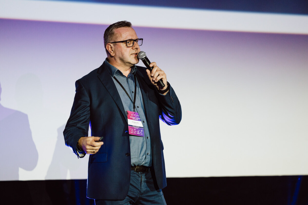 Mężczyzna w garniturze i okularach mówi do mikrofonu podczas prezentacji na scenie, trzymając kliker. Nosi plakietkę konferencyjną i stoi przed ekranem projekcyjnym z fioletowym i niebieskim oświetleniem.
