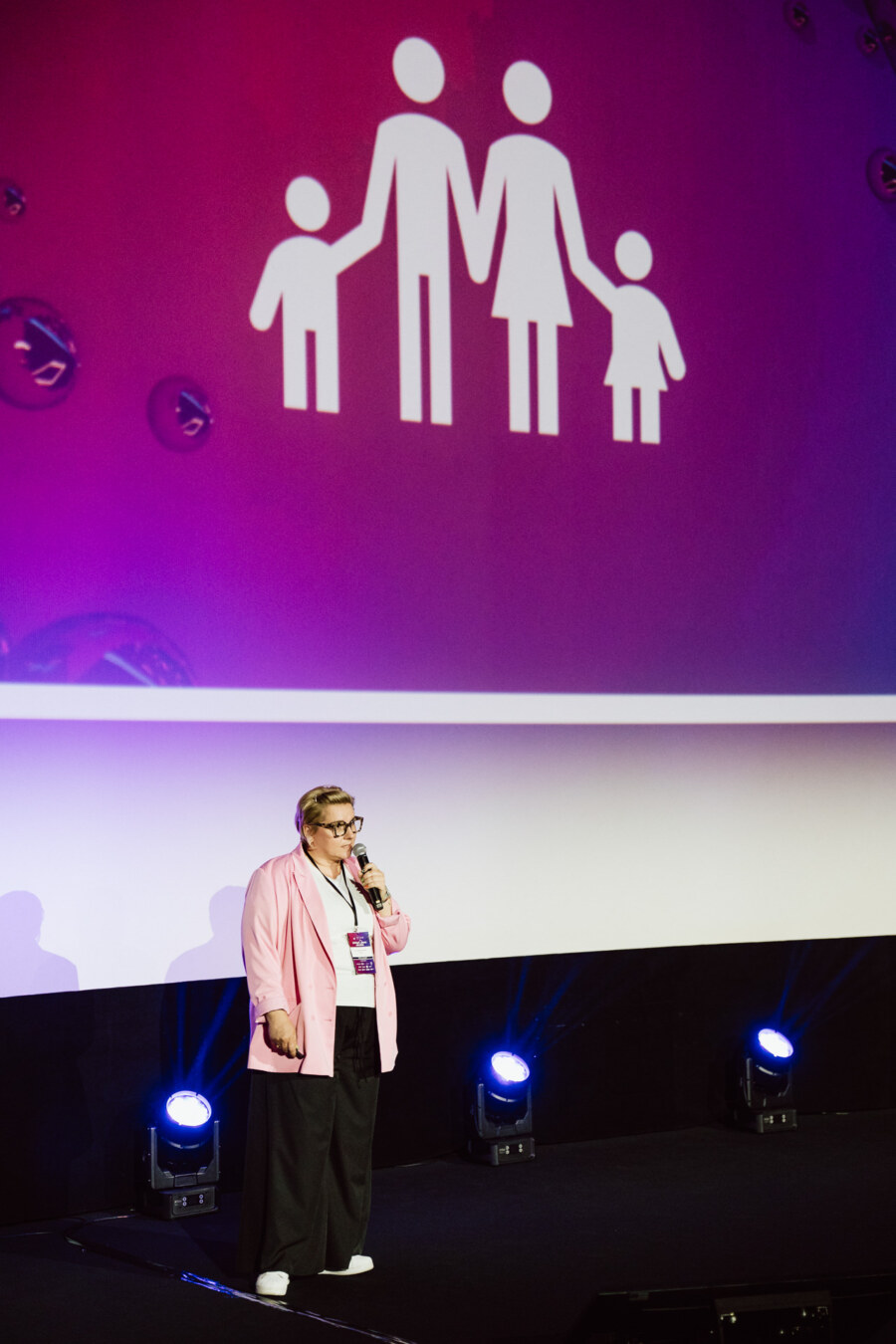 Osoba w różowej marynarce przemawia na scenie z mikrofonem, stojąc przed dużym ekranem wyświetlającym białą ikonę czteroosobowej rodziny na fioletowym tle.