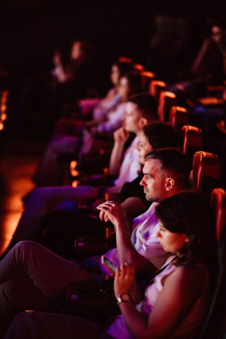Grupa ludzi siedząca w słabo oświetlonym teatrze, uważnie oglądająca przedstawienie lub film. Ich twarze są delikatnie oświetlone przez ekran i siedzą na czerwonych krzesłach w rzędzie.