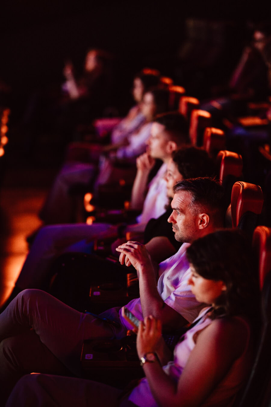 Eine Gruppe von Menschen, die in einem schwach beleuchteten Theater sitzen und sich aufmerksam eine Aufführung oder einen Film ansehen. Ihre Gesichter werden von der Leinwand sanft beleuchtet und sie sitzen auf roten Stühlen in einer Reihe. 