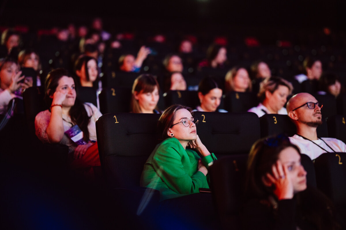 Grupa ludzi siedzi w fotelach teatralnych, uważnie obserwując coś poza kamerą. Większość wydaje się być zaangażowana lub zamyślona; kobieta w zielonej kurtce siedzi pośrodku, opierając brodę na dłoni.