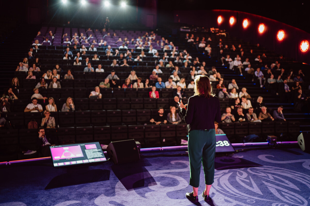 Na scenie stoi osoba przemawiająca do dużej publiczności siedzącej w teatrze. Jasne światła sceniczne świecą w dół, a dwa ekrany wyświetlają wizualizacje w pobliżu mówcy. Publiczność stoi twarzą do sceny, słuchając uważnie.