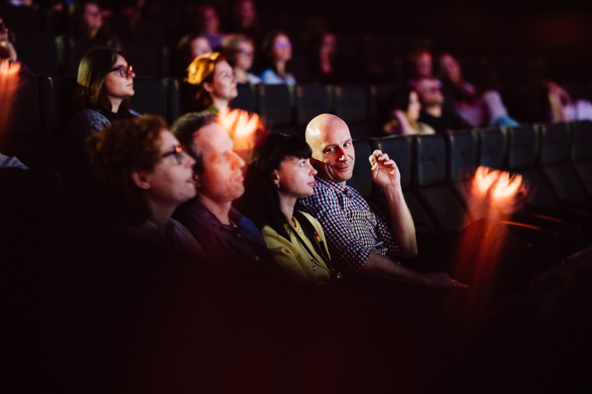 Widzowie siedzą w zaciemnionym teatrze i oglądają wydarzenie. Jedna osoba w pobliżu centrum patrzy w stronę kamery i gestykuluje, podczas gdy inni skupiają się na scenie. Miejsca za nimi są wypełnione uważnymi widzami.