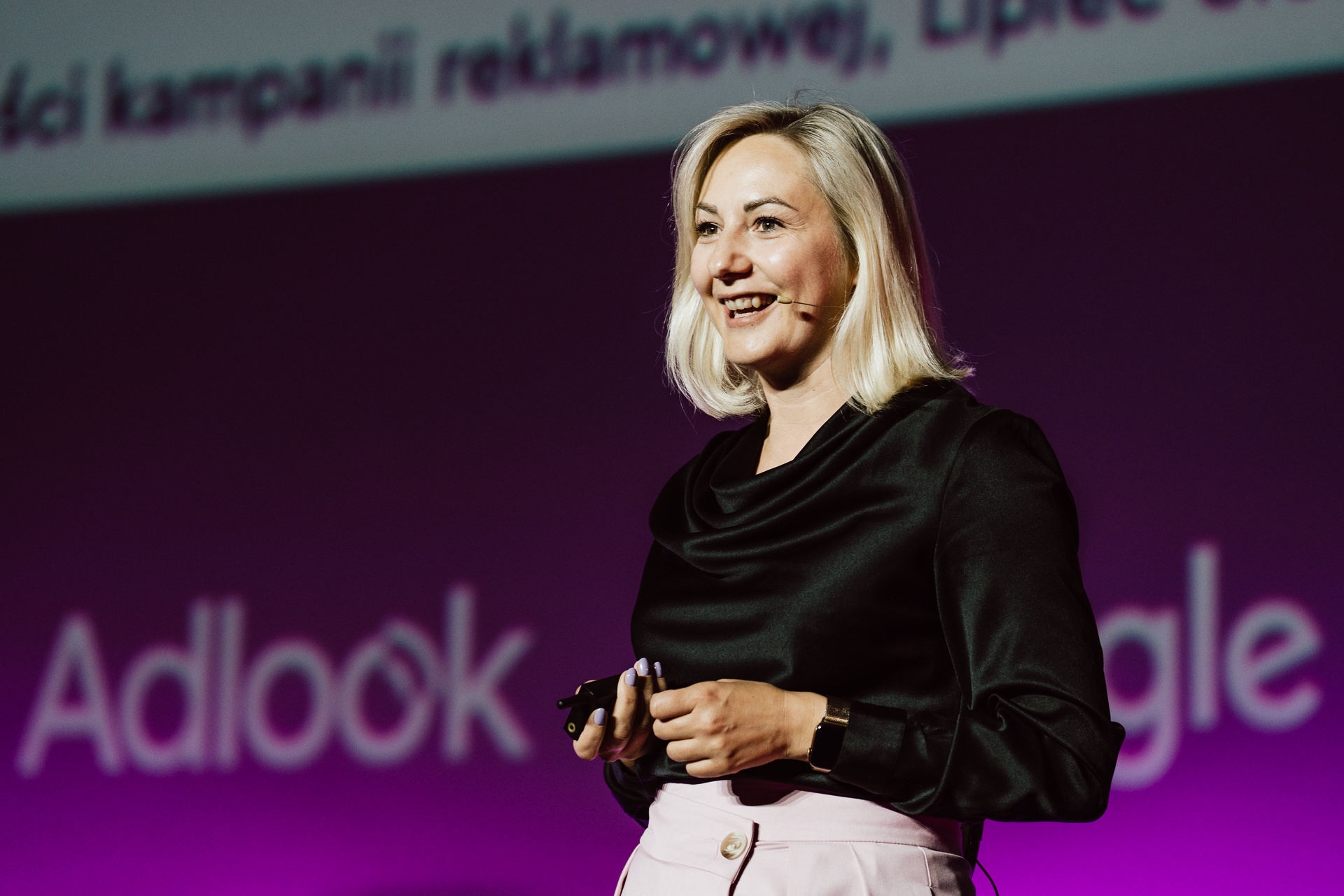 Fotorelacja z konferencji - Kobieta o blond włosach, ubrana w czarną bluzkę i jasne spodnie, uśmiecha się, przemawiając na scenie z mikrofonem w słuchawkach. Fioletowe tło wyświetla rozmazany tekst i logo, w tym "Adlook" i "Google".