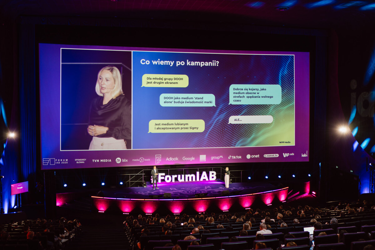 Kobieta stoi na scenie przed dużym ekranem wyświetlającym prezentację w języku polskim dla publiczności w słabo oświetlonym audytorium w Forum IAB. Scena jest oświetlona fioletowymi światłami.