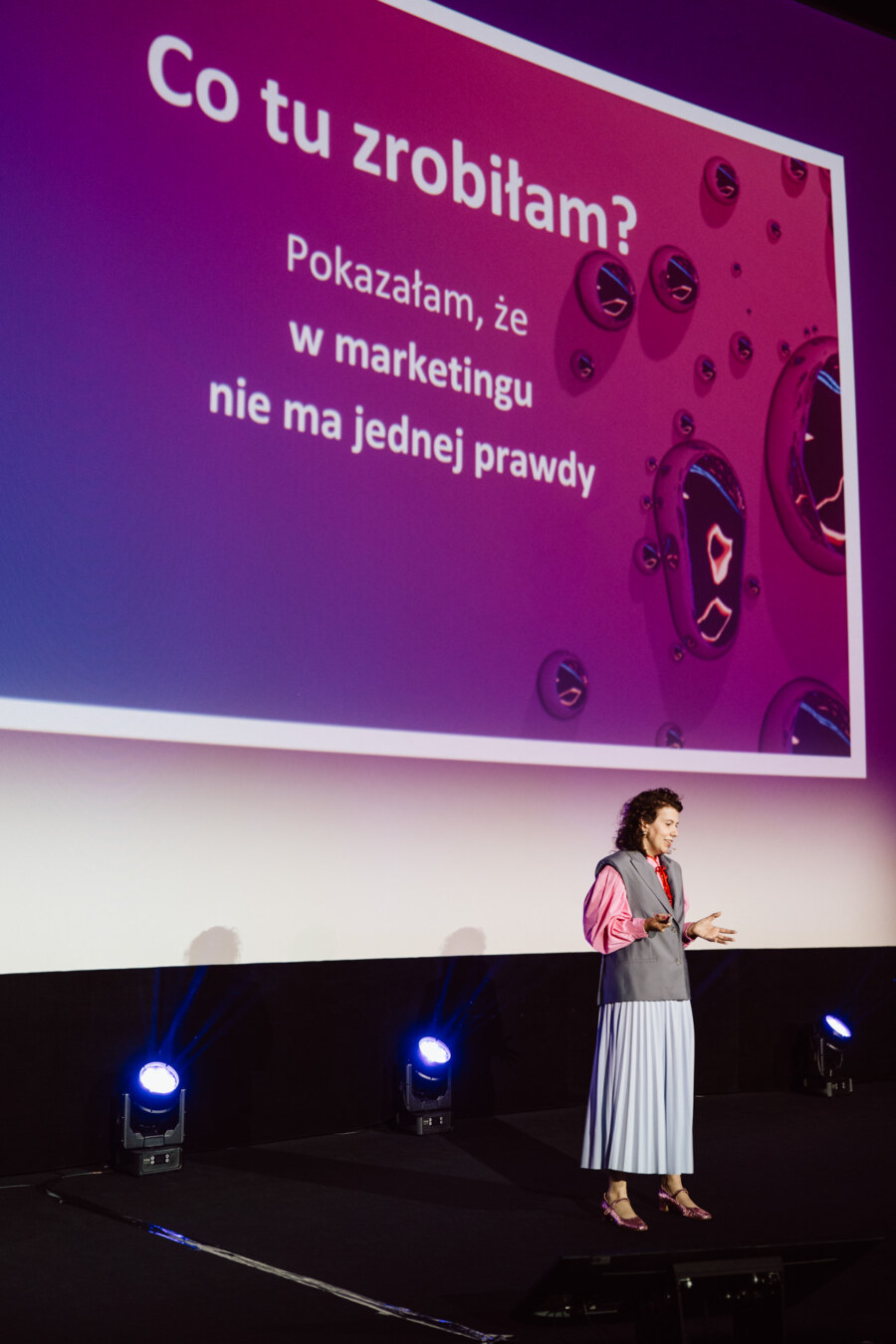 Kobieta stoi na scenie, prowadząc prezentację przed dużym ekranem z polskim tekstem: "Co tu zrobiłam? Pokazałam, że w marketingu nie ma jednej prawdy". Niebieskie i fioletowe oświetlenie oświetla scenę.