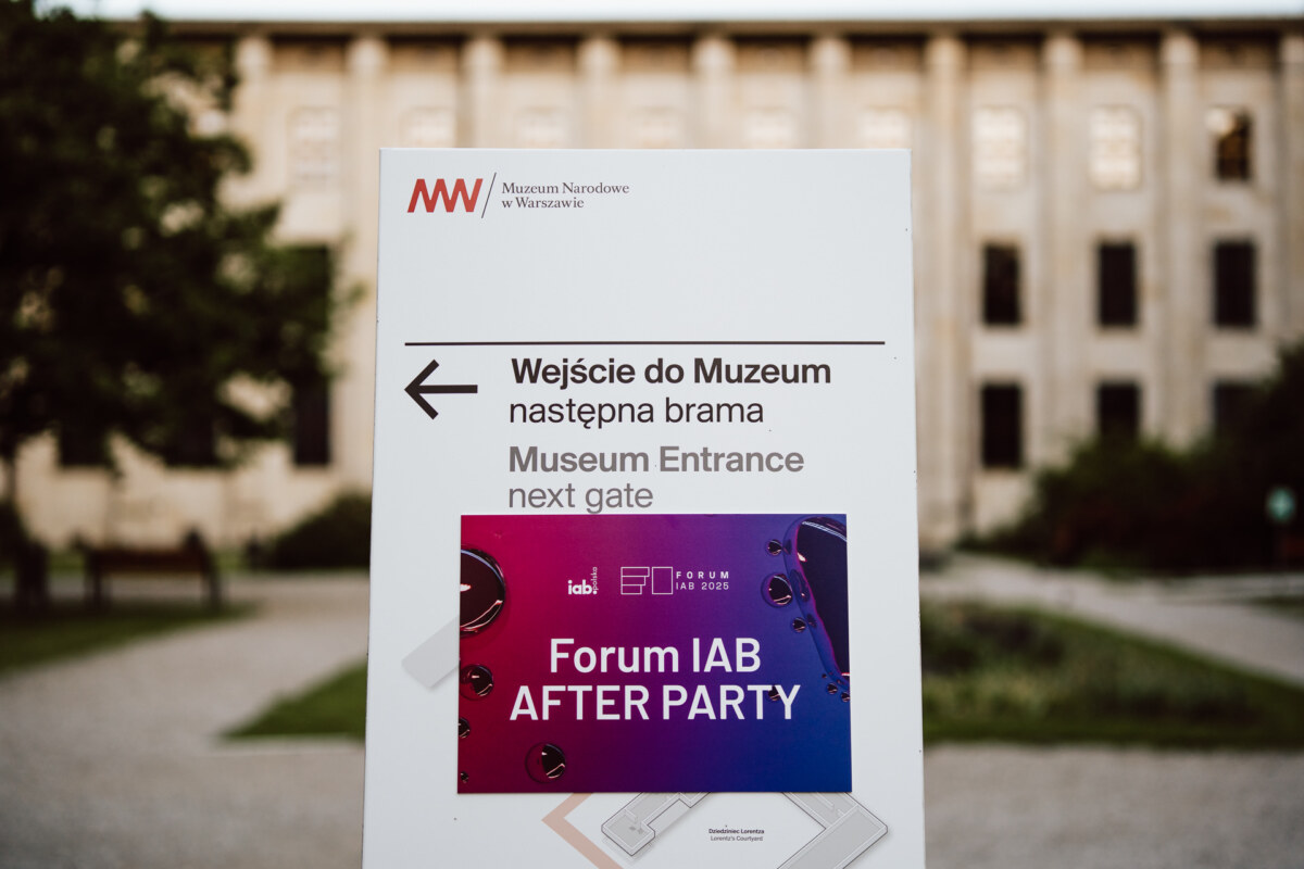 Znak przed budynkiem z napisem "Wejście do muzeum obok bramy" w języku polskim i angielskim, z fioletowym plakatem "Forum IAB AFTER PARTY" zakrywającym część znaku. W tle widać drzewa i duży budynek.