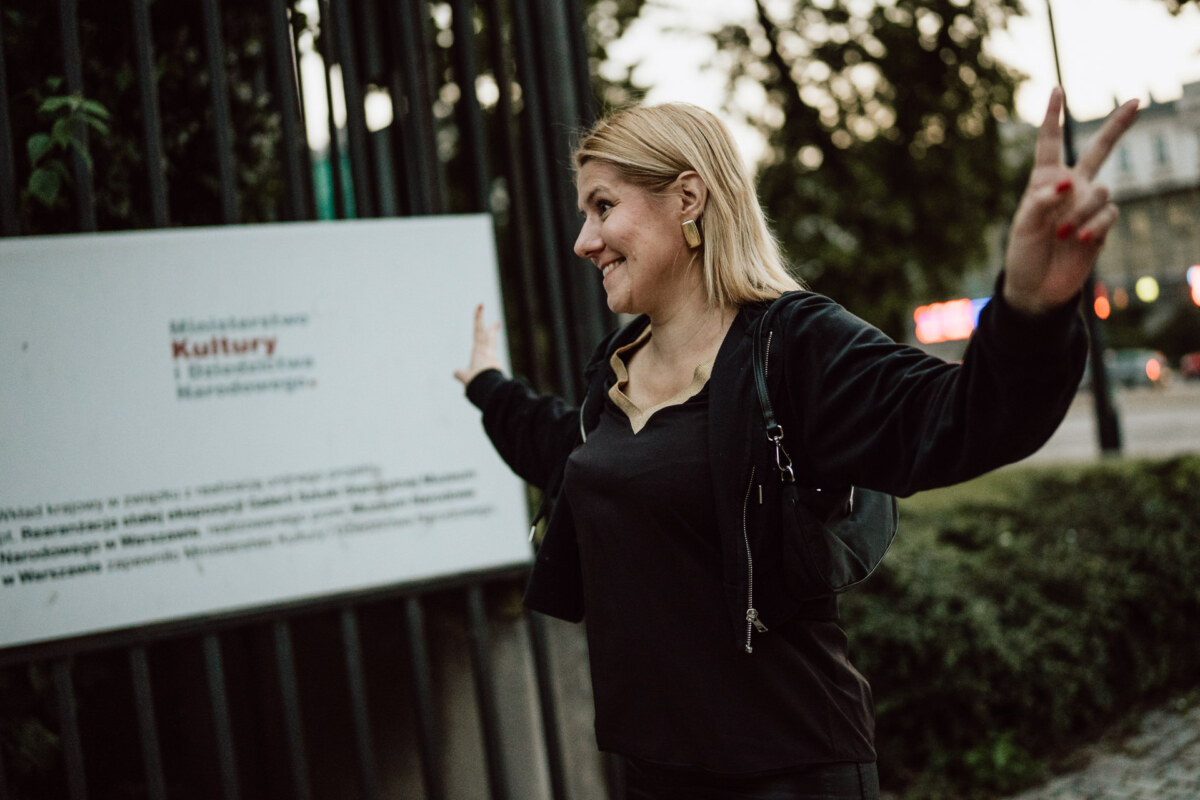 Uśmiechnięta kobieta o blond włosach stoi na zewnątrz w pobliżu znaku, trzymając obie ręce w znakach pokoju. Ma na sobie czarną kurtkę i patrzy w bok. W tle widoczne są drzewa i budynki.