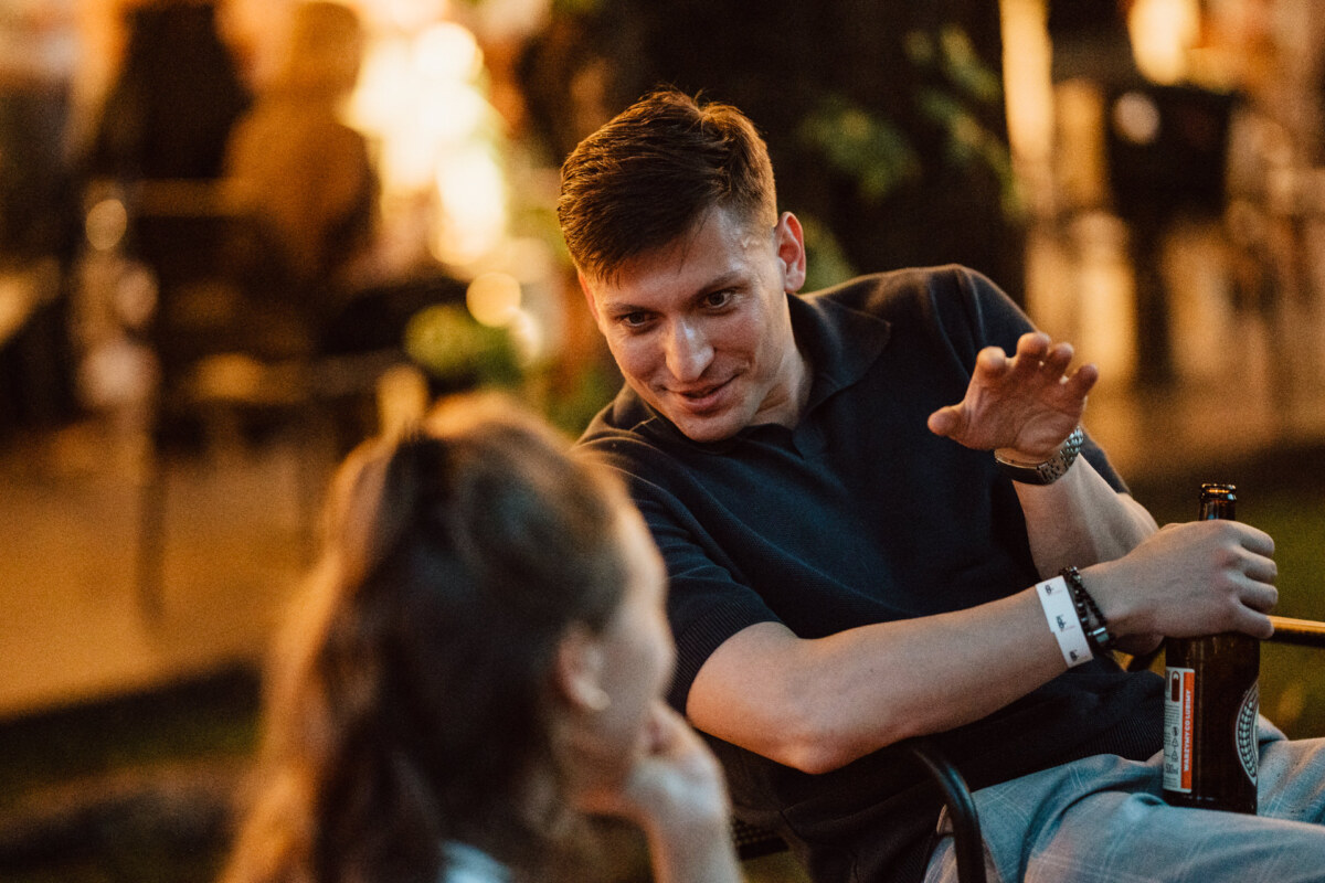Mężczyzna trzymający butelkę piwa żywo gestykuluje podczas rozmowy z kobietą na wieczornym spotkaniu na świeżym powietrzu, z ciepłymi, rozmytymi światłami w tle.