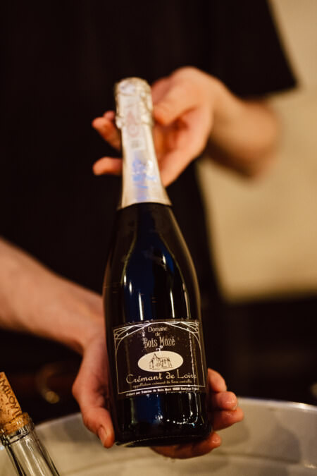 Eine Person hält eine Flasche Domaine Bois Mozé Crémant de Loire Sekt und präsentiert sie mit beiden Händen. Der Hintergrund ist unscharf und lenkt die Aufmerksamkeit auf die Flasche. 