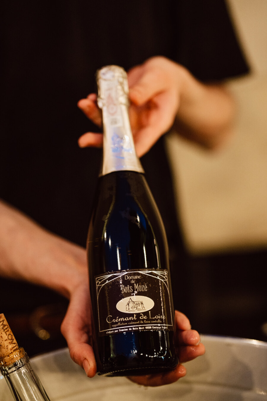 Osoba trzyma butelkę wina musującego Domaine Bois Mozé Crémant de Loire, prezentując ją obiema rękami. Tło jest rozmyte, przyciągając uwagę do butelki.