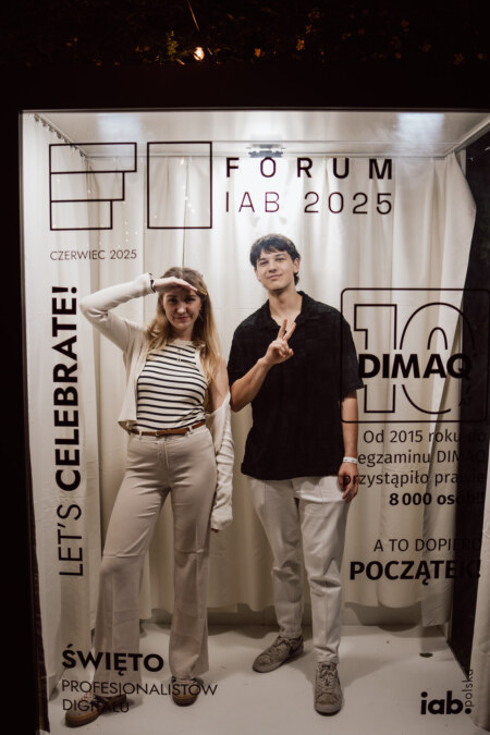 Kobieta i mężczyzna pozują i uśmiechają się w budce fotograficznej z napisem "FORUM IAB 2025" i różnymi polskimi tekstami nadrukowanymi na szybie. Kobieta wskazuje w górę, a mężczyzna salutuje. Jako tło wiszą białe zasłony.