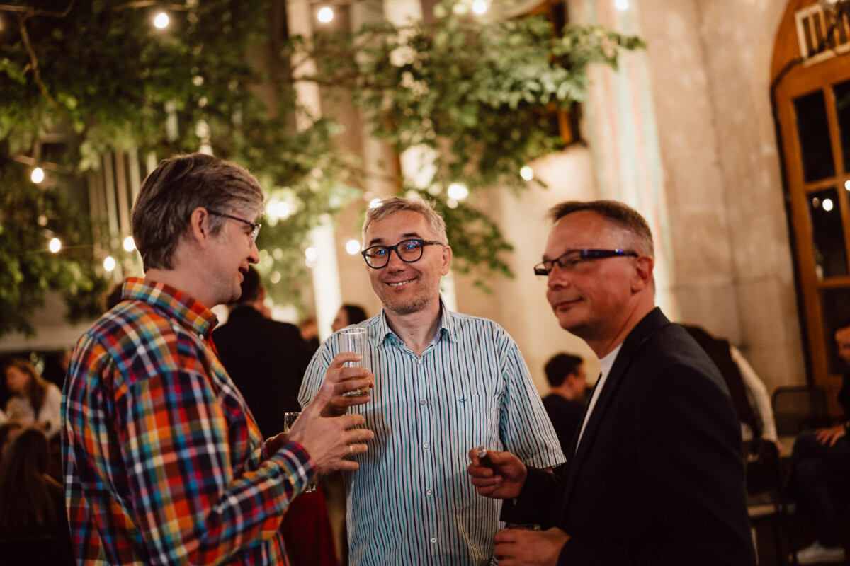 Trzech mężczyzn stoi razem na wieczornym spotkaniu na świeżym powietrzu, rozmawiając i trzymając drinki. Światła sznurkowe i zieleń tworzą ciepłą, świąteczną atmosferę w tle.
