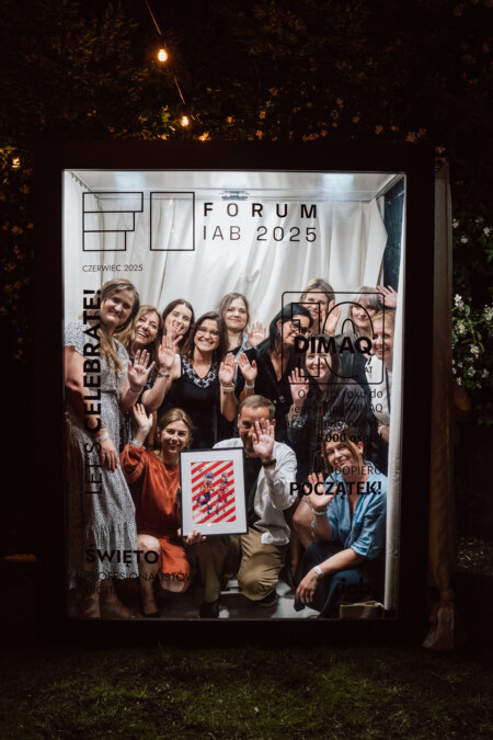 Grupa ludzi pozuje i uśmiecha się wewnątrz dużego, oświetlonego szklanego pudełka w nocy, trzymając kolorowy plakat i machając. Pudełko jest ozdobione różnymi polskimi słowami i grafikami dla "Forum IAB 2025".