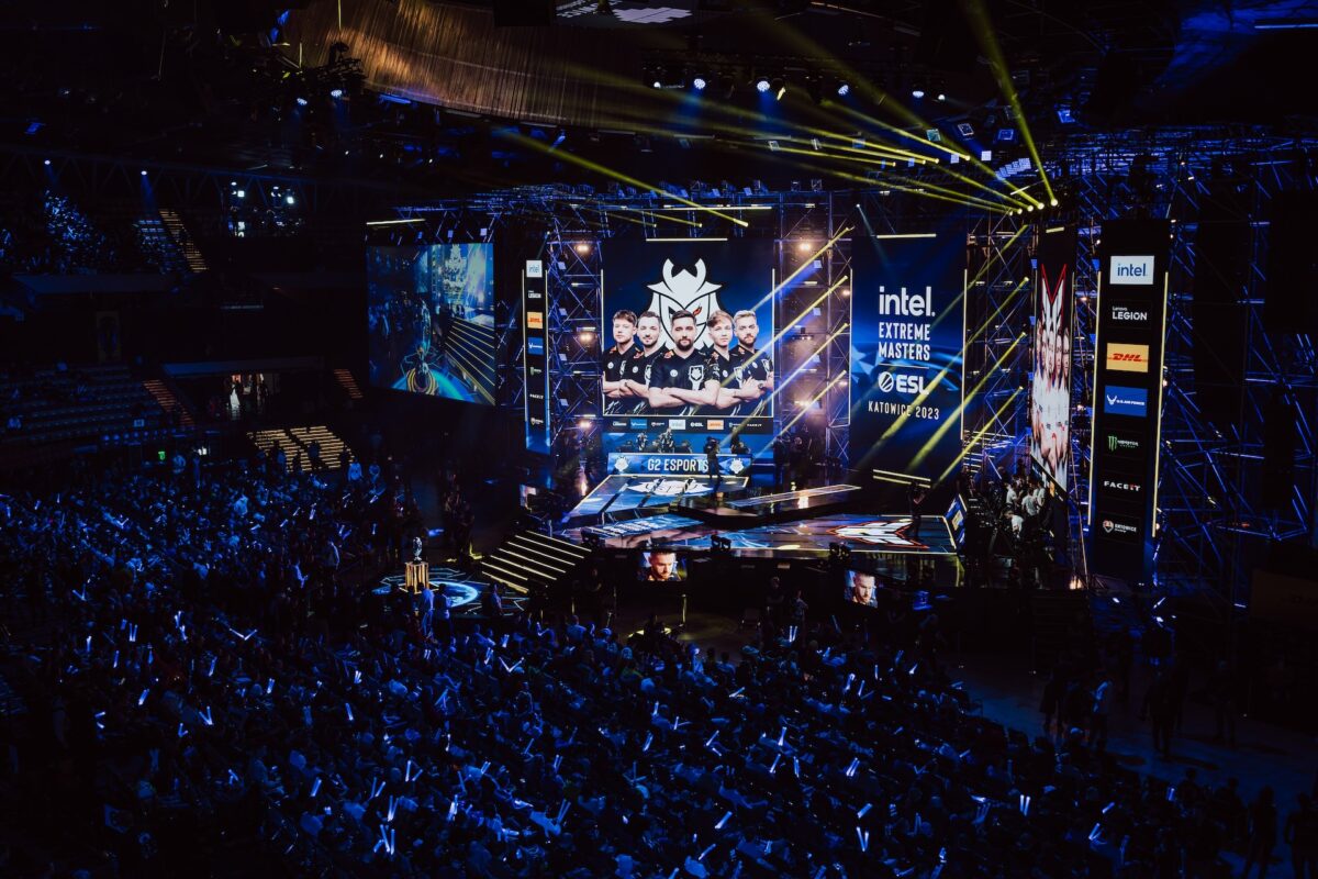 Duży tłum ogląda turniej e-sportowy na ciemnej arenie z niebieskimi światłami i świecącymi pałeczkami. Fotograf na imprezie pstryka zdjęcia, podczas gdy gigantyczny ekran wyświetla skład profesjonalnej drużyny i logo sponsorów Intela, ESL i innych na scenie.