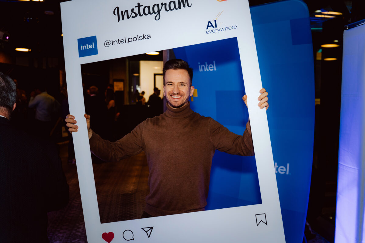 Mężczyzna w brązowym swetrze z golfem uśmiecha się, trzymając dużą ramkę na zdjęcia o tematyce Instagrama, uchwyconą przez fotografa na spotkaniu pod marką Intel i AI.