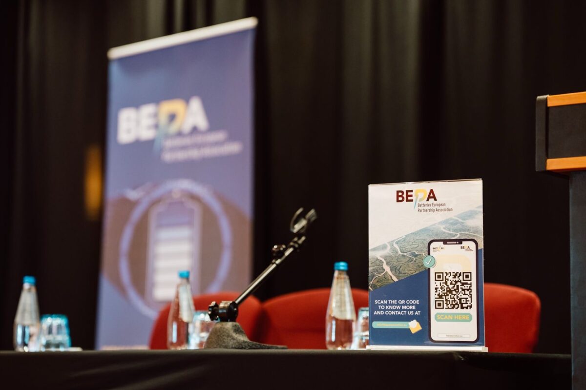 Stół konferencyjny z mikrofonami, butelkami wody i znakiem wydarzenia BEPA wyświetlającym kod QR; niewyraźny baner BEPA stoi w tle na tle czarnych zasłon - uchwycony przez profesjonalnego fotografa na wydarzeniu.